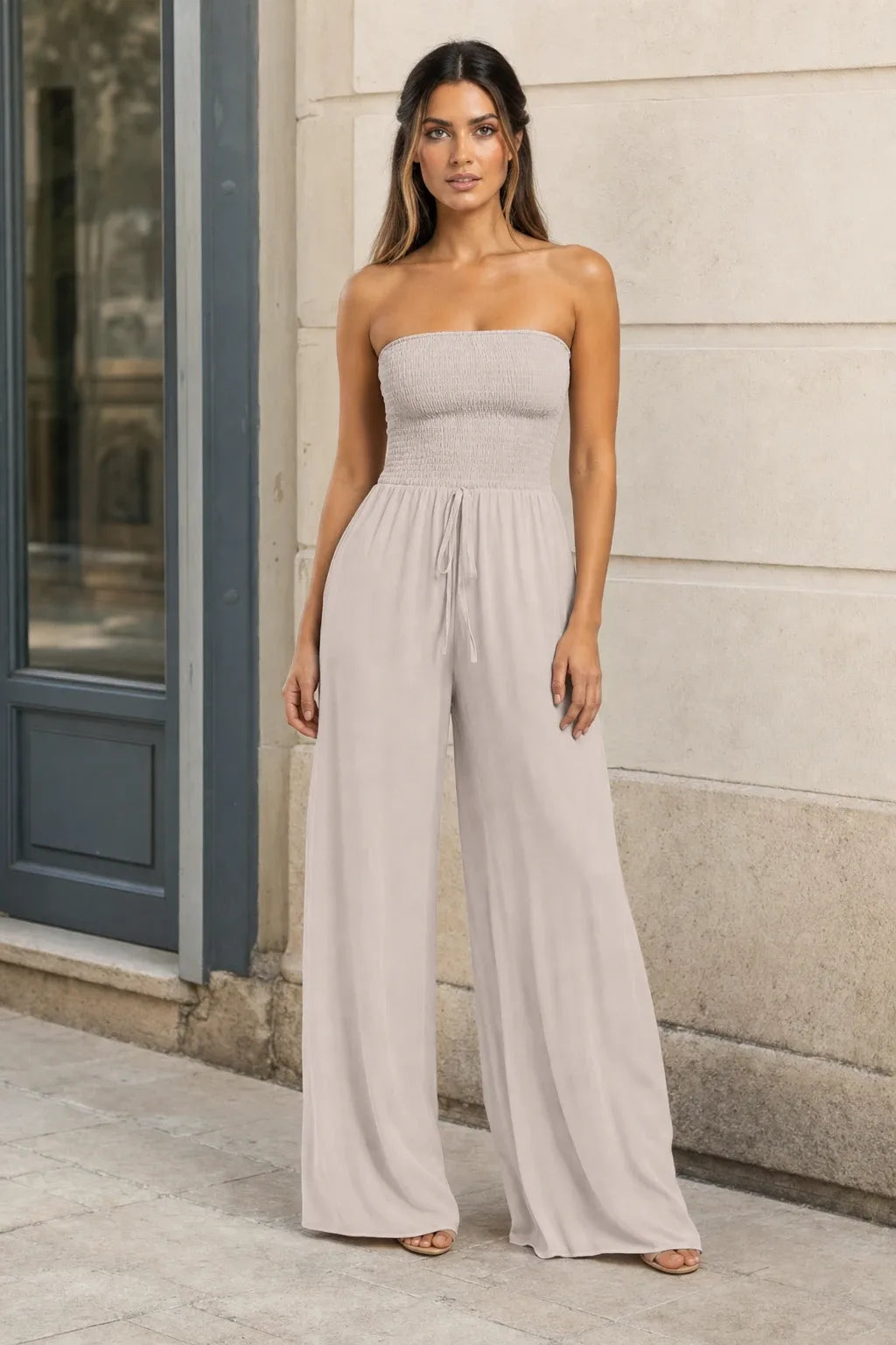 Beige smocked wide leg jumpsuit on model in contrapposto stance, elastic bodice and wide-leg trousers