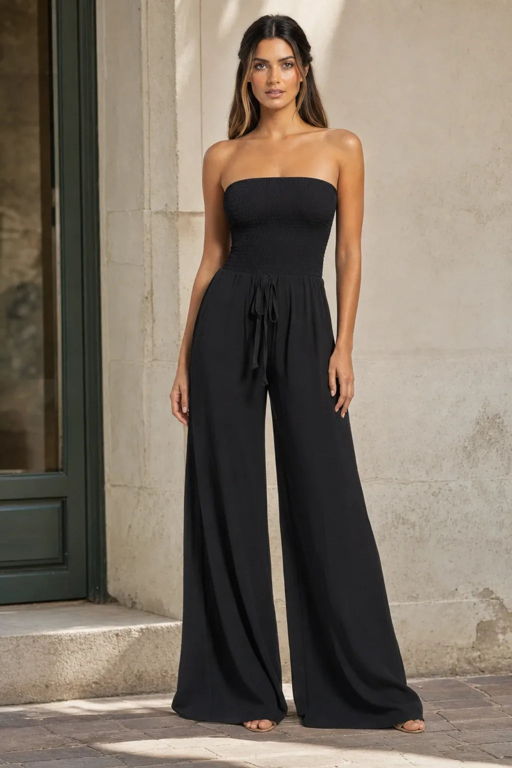 Black smocked wide leg jumpsuit on model in contrapposto stance, elastic bodice and wide-leg trousers